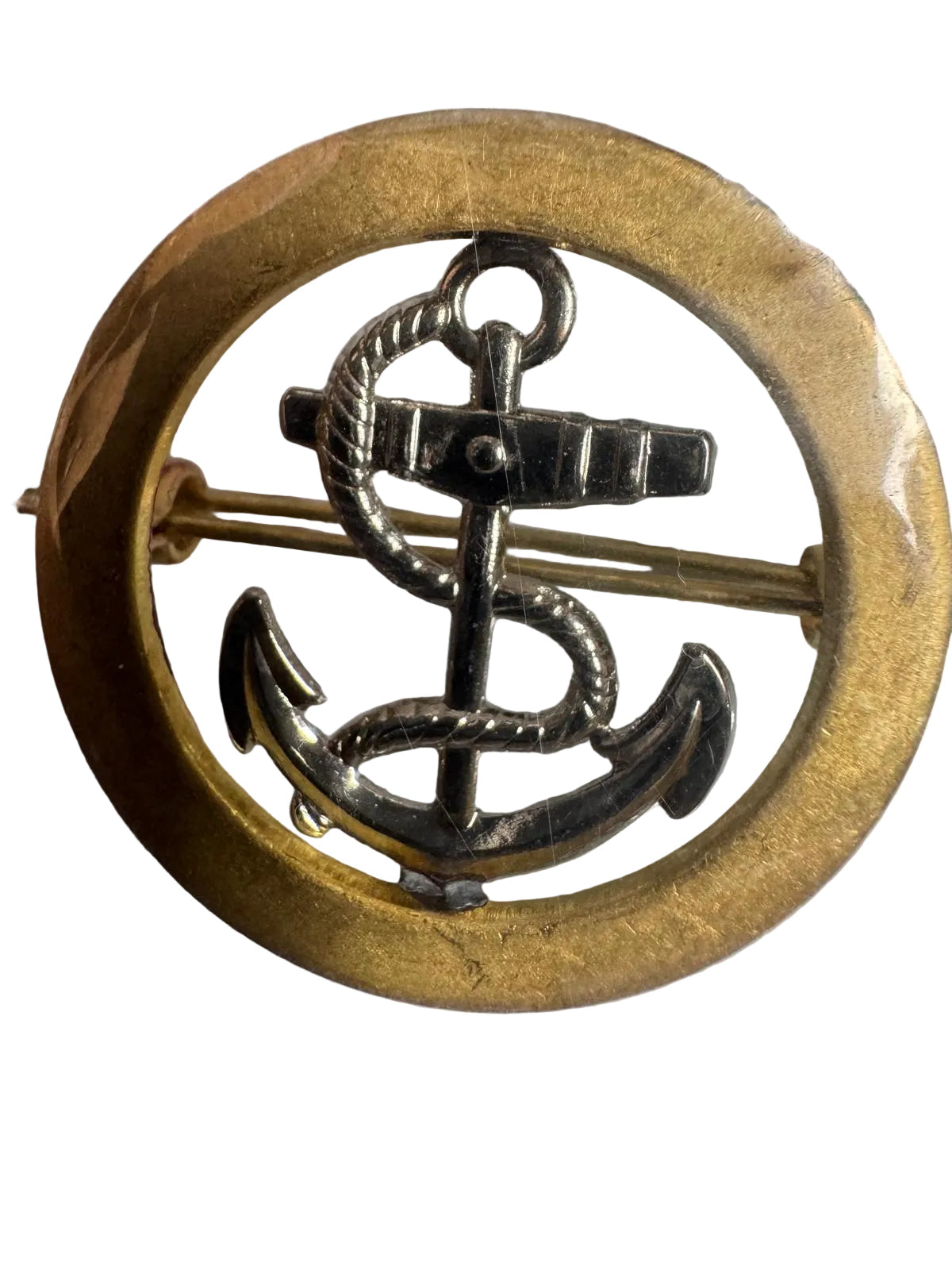 A circular gold-toned metal pin or badge featuring a silver-toned anchor and rope motif, photographed on a light surface with some plastic wrapping visible.