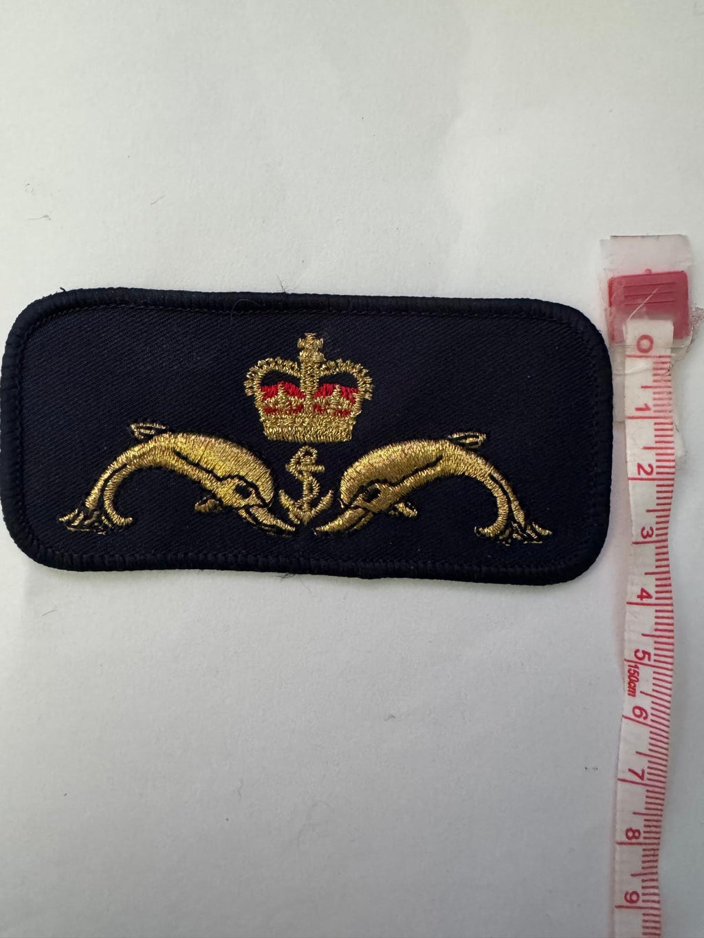 A rectangular embroidered patch with rounded corners, featuring two gold dolphins and a red-and-gold crown on a dark navy background. A pink measuring tape is placed above the patch to indicate size, set against a plain off-white surface.