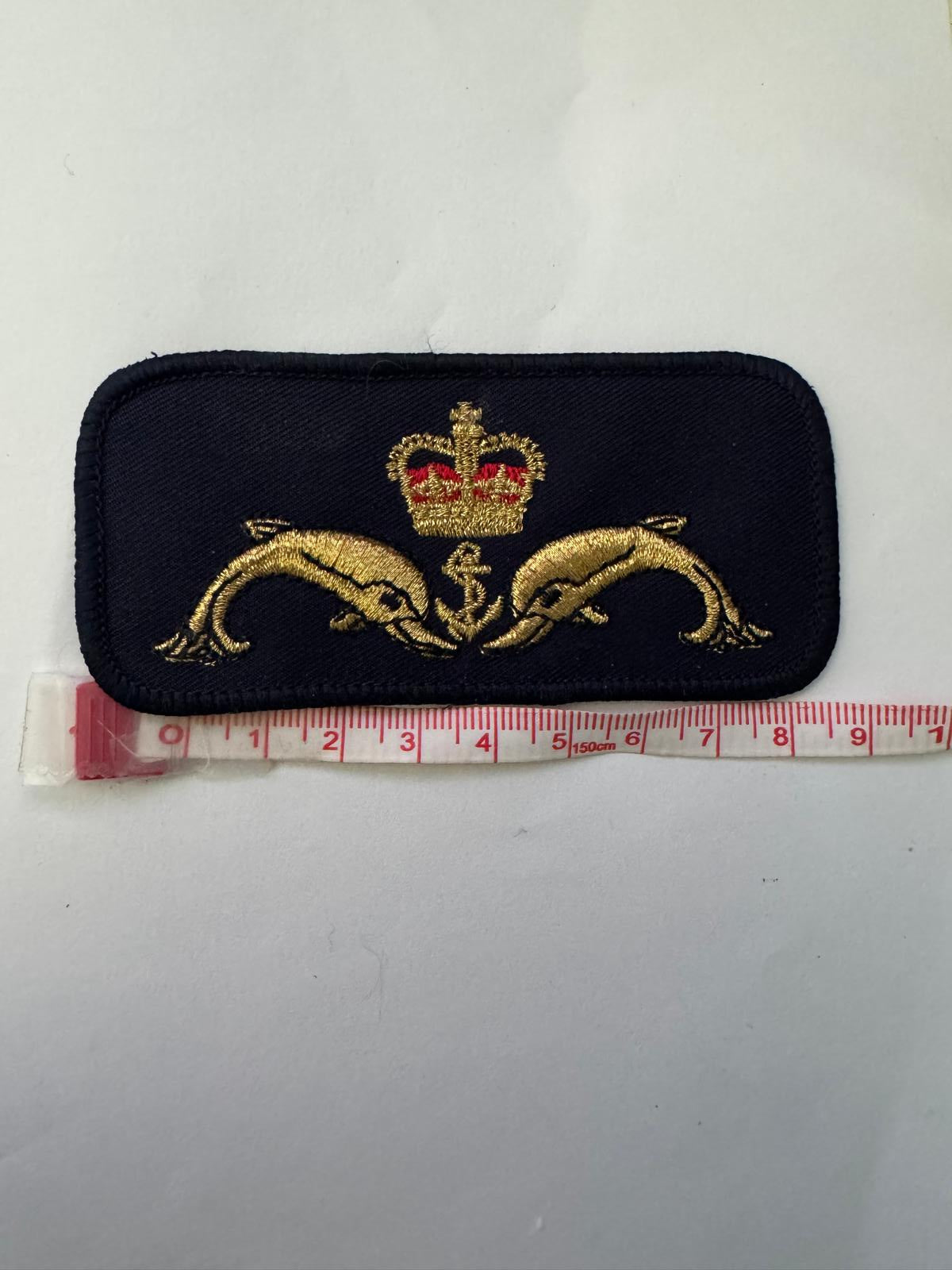 A small embroidered patch on a white background next to a measuring tape. The patch is dark navy blue with two gold dolphins and a gold crown with a red jewel.