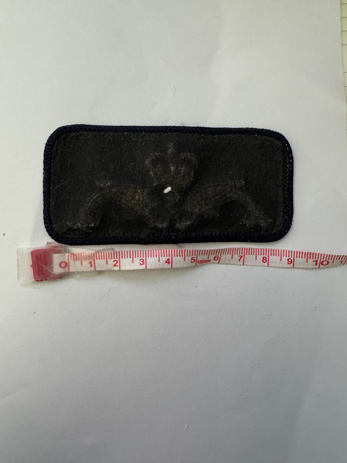 A black rectangular fabric pouch or case with a dark embroidered design, shown next to a red and white measuring tape on a white background.
