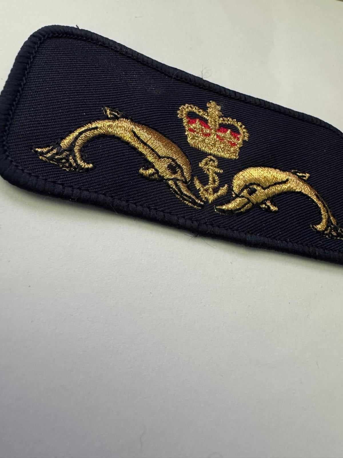 A close-up of a dark navy blue embroidered fabric patch featuring two gold dolphins and a small red and gold crown motif, placed on a light gray background.